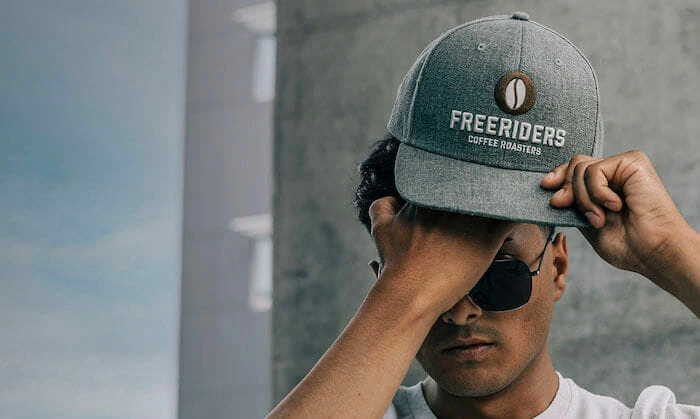 A heather gray snapback cap with a brown leather patch embroidered with "Freeriders Coffee Roasters," worn by a person adjusting the cap.