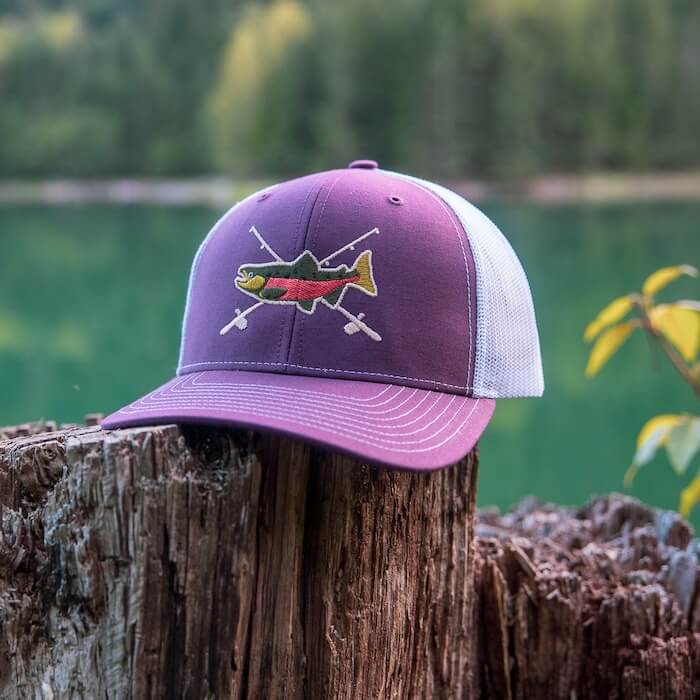 A purple and white trucker hat featuring an embroidered fish and paddles design, placed on a weathered tree stump near a lake.