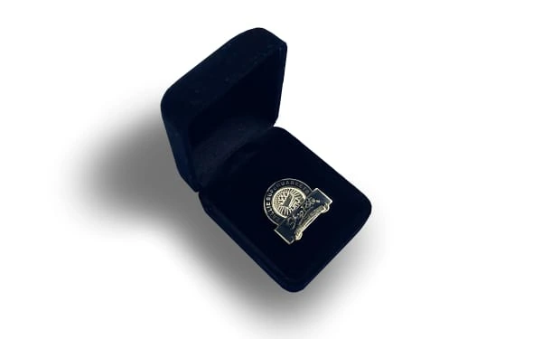 Gold lapel pin presented in an open black velvet box. The pin features an intricate engraved design with a circular emblem and banner detail.