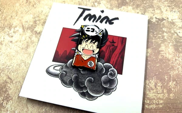 Custom enamel pin featuring a chibi-style anime character with a wide smile, wearing an orange martial arts uniform with a Japanese symbol on the front. The character is attached to a themed backing card displaying a red and black illustration of mountains and a cloudy sky with stylized text at the top.