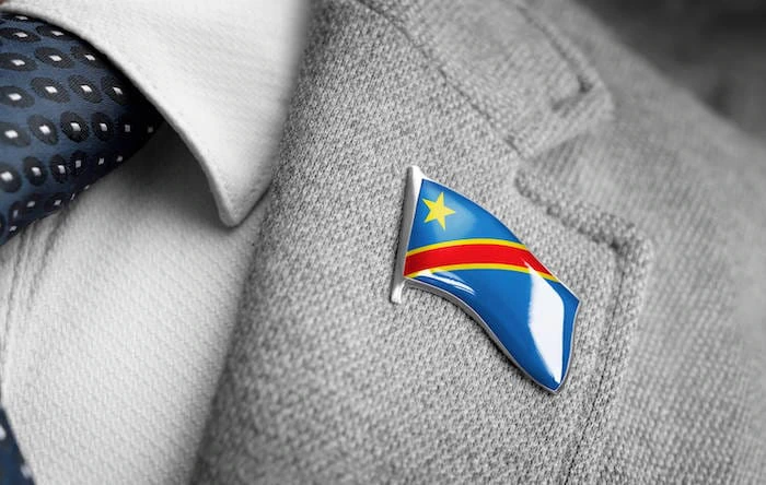 Close-up of a Democratic Republic of the Congo flag lapel pin attached to the lapel of a gray suit jacket. The pin features a wavy enamel finish with a yellow star, red diagonal stripe, and blue background.