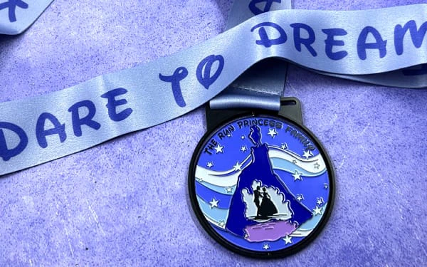 Custom racing medal with a princess theme, featuring a silhouette of a princess in a flowing gown with a castle scene inside. The background includes stars and flowing ribbons in shades of blue and purple. The medal is attached to a satin ribbon that reads 'DARE TO DREAM' in bold letters.