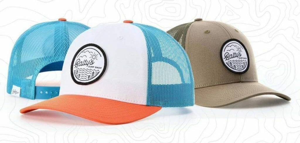 Three custom trucker hats displayed side by side. The first hat has a white front panel, blue mesh back, and an orange brim, while the second is fully khaki-colored. Both feature a circular 'Salty's Surf Shop' embroidered patch with a wave and sun design. The third hat, partially visible, shows a blue mesh back with an adjustable snap closure.