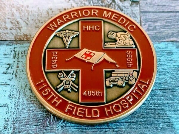 Round military challenge coin with a red and gold design, representing the 115th Field Hospital. The coin features a central red cross with a field tent and four quadrants containing medical and military symbols, including a caduceus, a combat medic, a transport vehicle, and a medical insignia. The text 'Warrior Medic' is inscribed at the top, with '115th Field Hospital' at the bottom, along with unit designations 'HHC,' '643rd,' '565th,' and '485th.