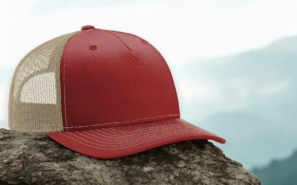 Red and tan trucker hat placed on a rock with a misty mountain landscape in the background. The hat has a solid red front panel, a curved brim, and a breathable tan mesh back, giving it a rugged and outdoorsy look.