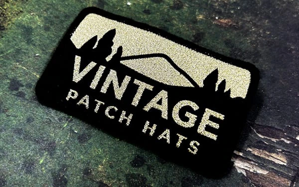 Rectangular woven patch with a black background and gold embroidery, featuring a mountain landscape silhouette and the text 'VINTAGE PATCH HATS.' The patch is displayed on a textured, rustic surface.